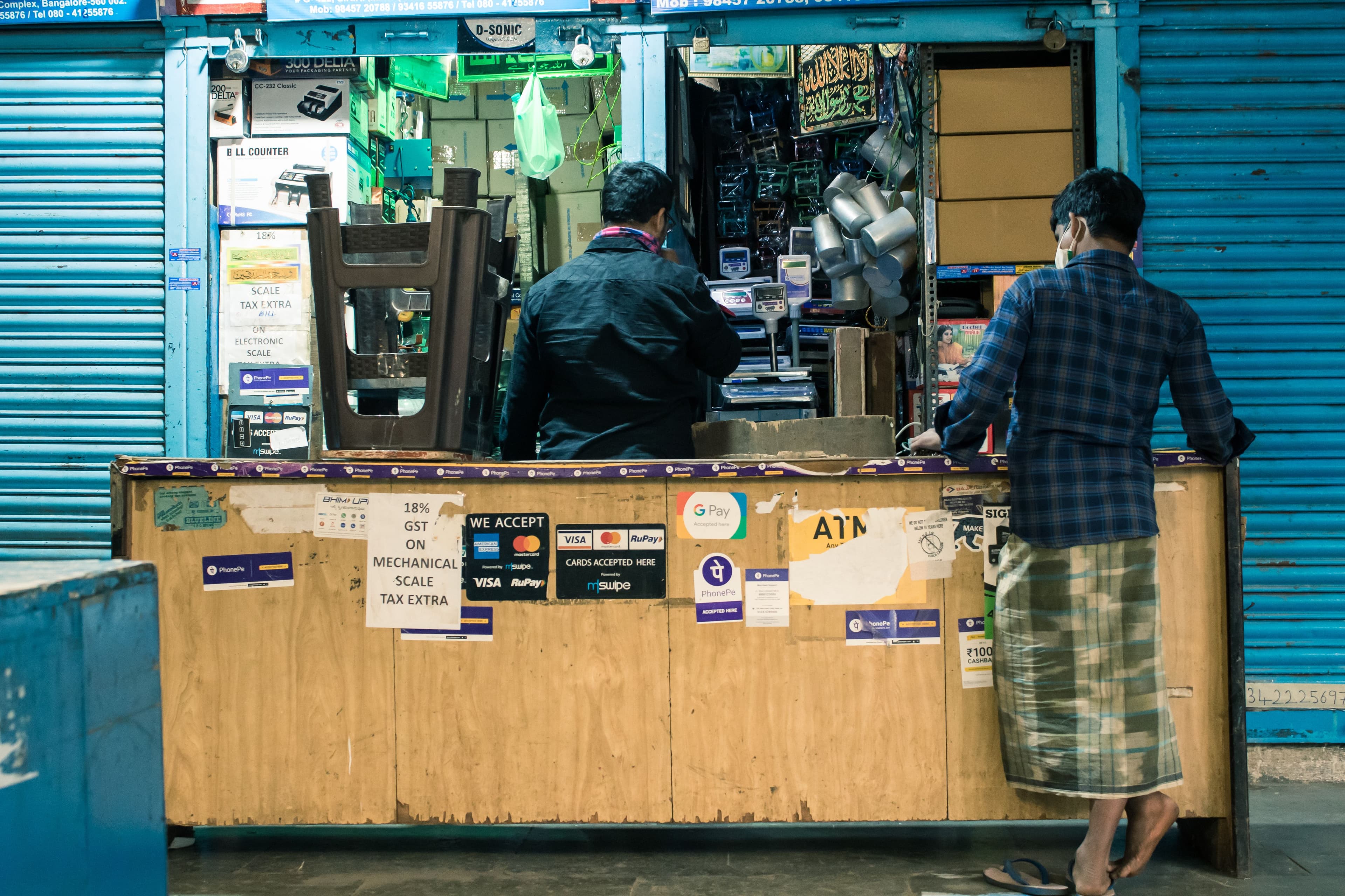 Two men at a shop counter in a Bengaluru marketplace, with GPay, card and UPI payment-acceptance signs taped to the counter