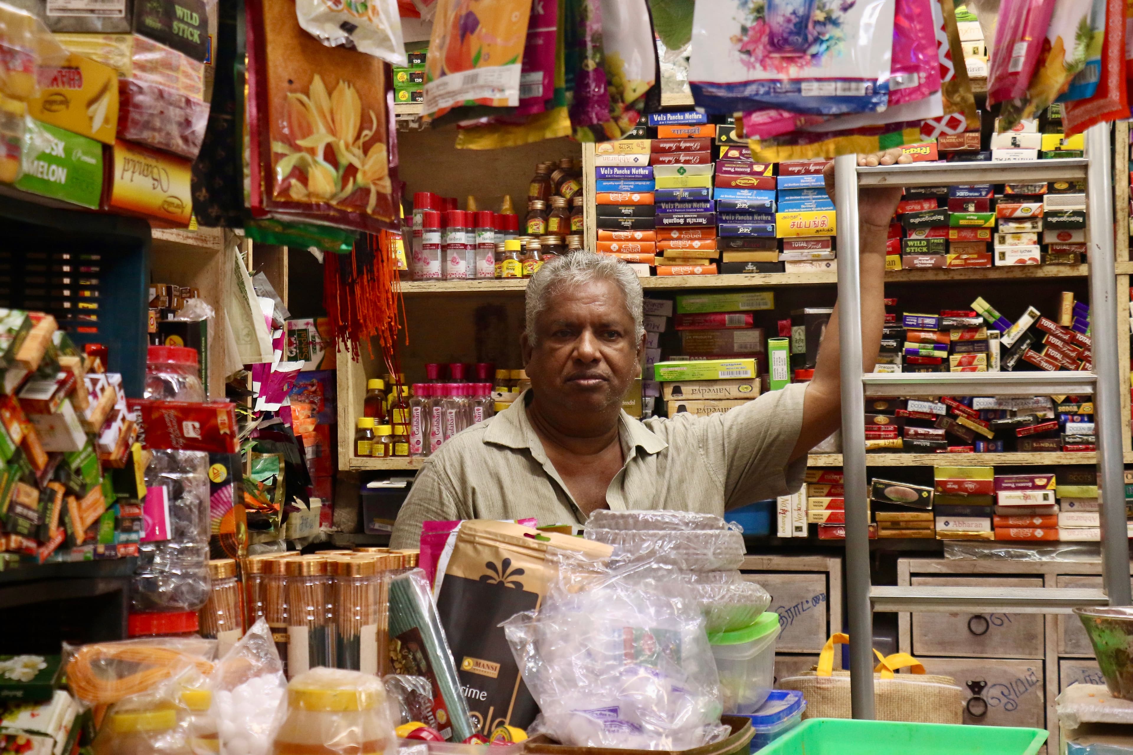 A middle-aged man working behind the counter of a small, densely stocked Indian grocery store