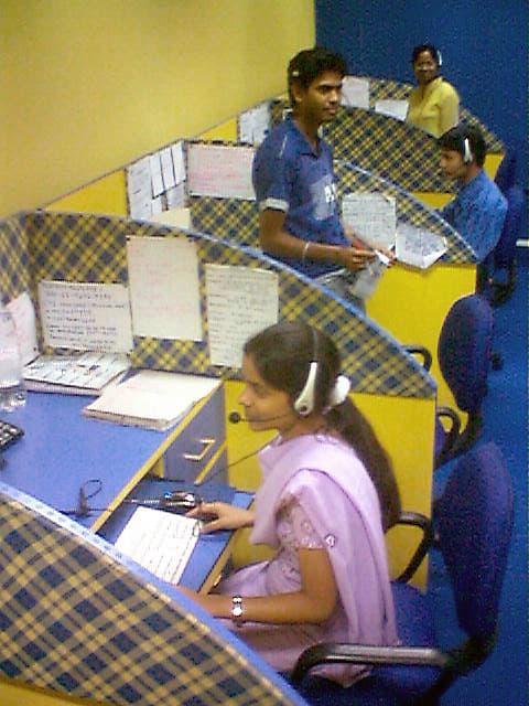 Customer-service agents wearing headsets at cubicle desks in an Indian call center, with a woman in a pink sari at the nearest desk